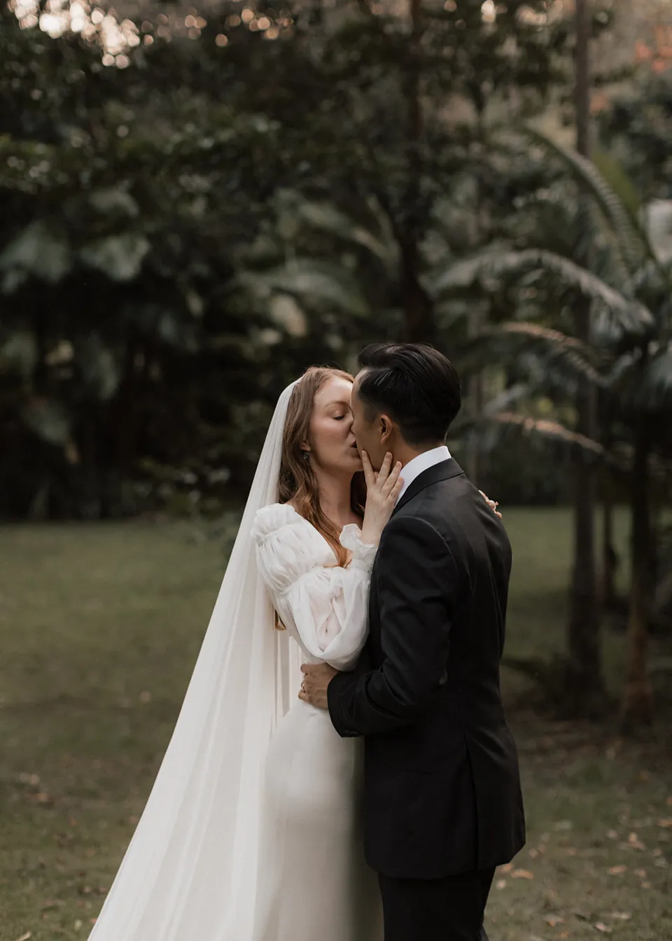 Elyse lifting her hand to Jonathan's face as they stand together. At Fig Tree Villa in Ewingsdale, Byron Bay, for an Elopement Collective elopement. Photograph by Bulb Creative.