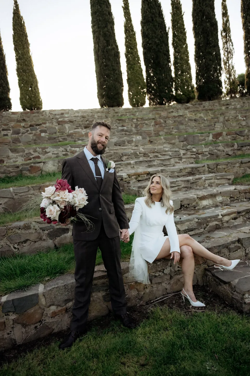 Russell holding the bouquet by The Romantics while Kaitlyn sits laughing on the stone amphitheatre steps. At Piermont Retreat on Tasmania’s east coast, for an Elopement Collective elopement. Photograph by Omar of By The Wilde.