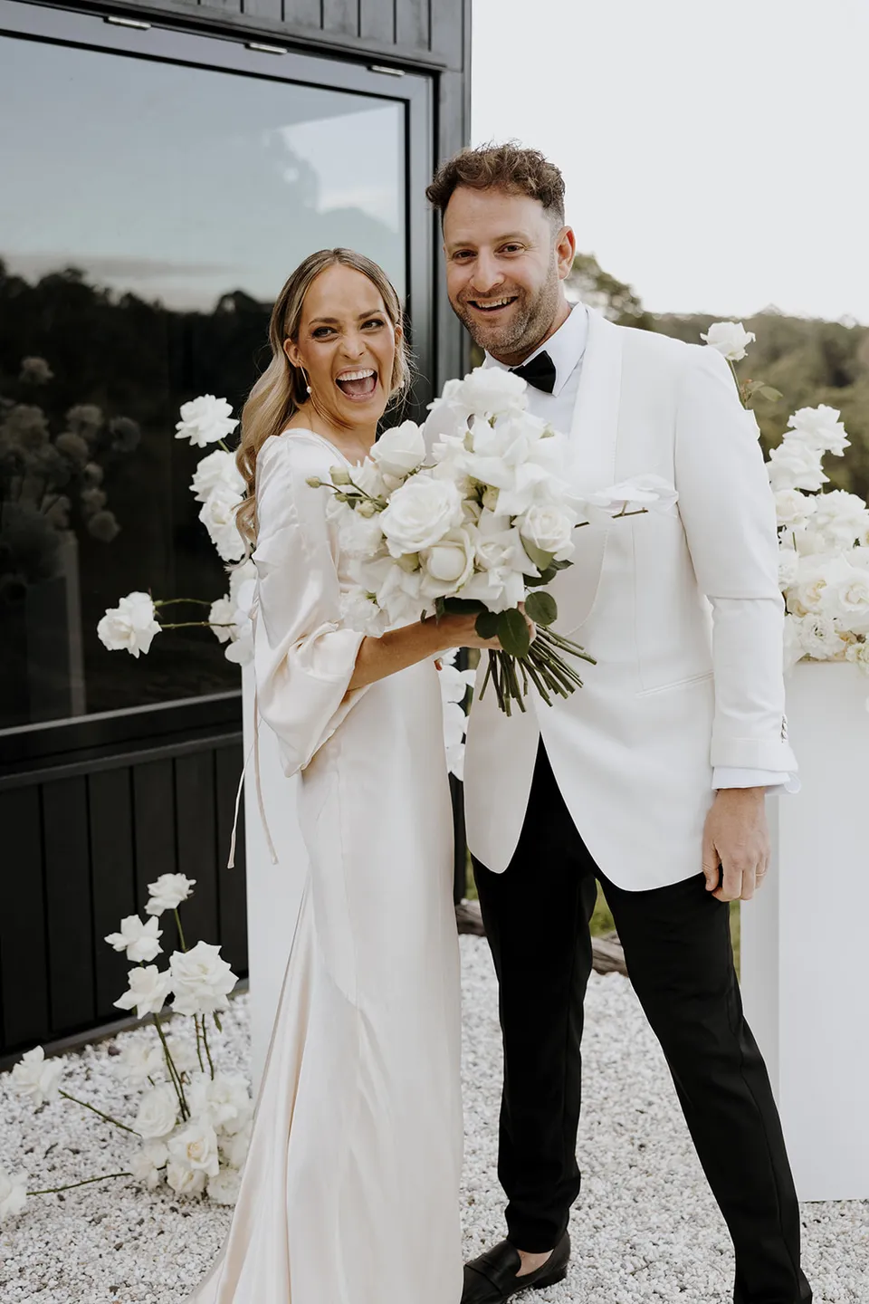 Gabrielle and Greg smiling together with her white bouquet by Mrs Gibbons beside the cabin after their ceremony, at Roundhill Retreat in Obi Obi, Sunshine Coast Hinterland, Queensland, for an Elopement Collective elopement. Photo by George Bowden.