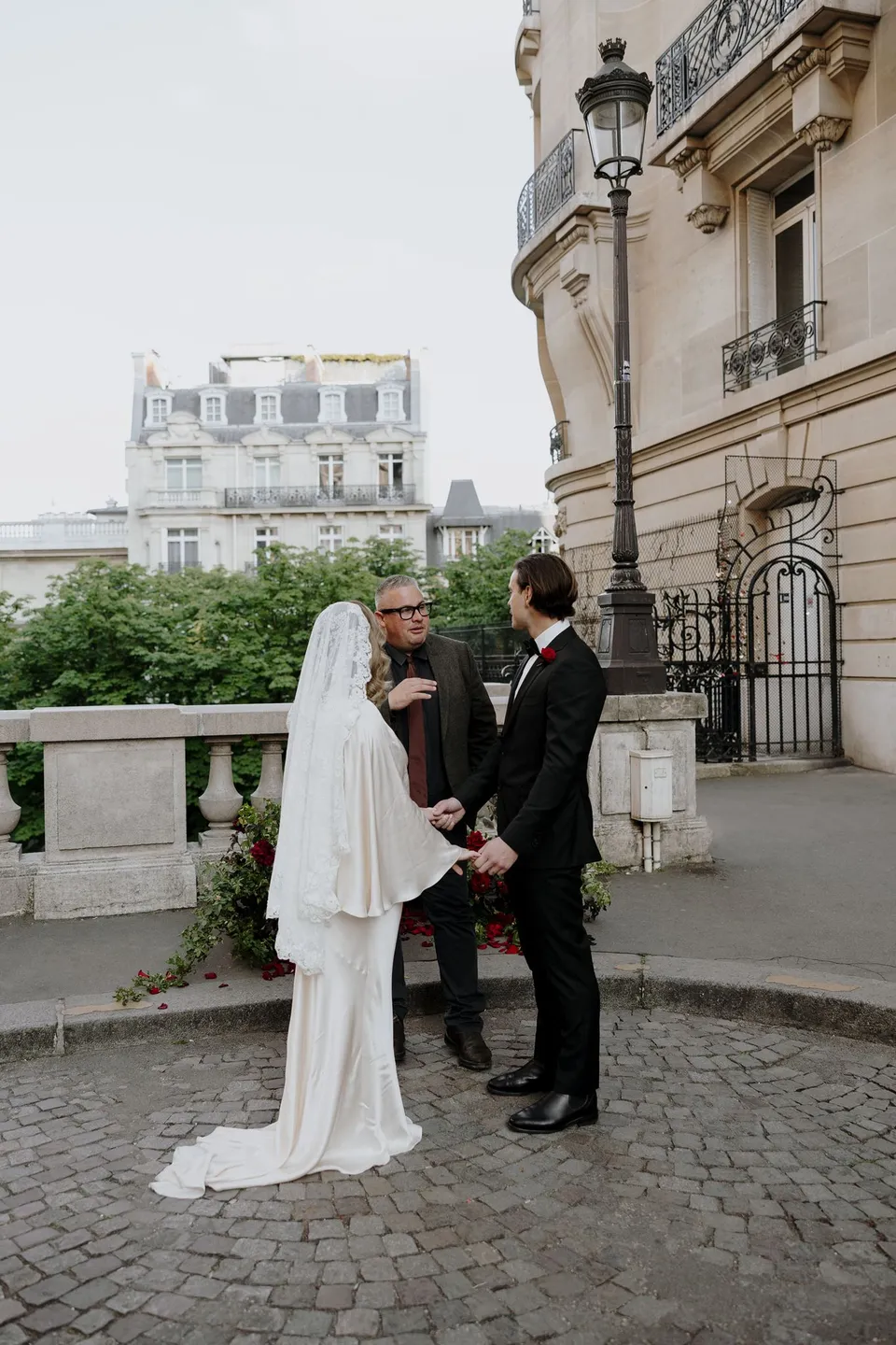 Celebrant Josh Withers standing with Kara and Samuel as they face each other beside the stone balustrade, at Avenue de Camoëns in Paris, France, during an Elopement Collective elopement. Photograph by George Bowden.