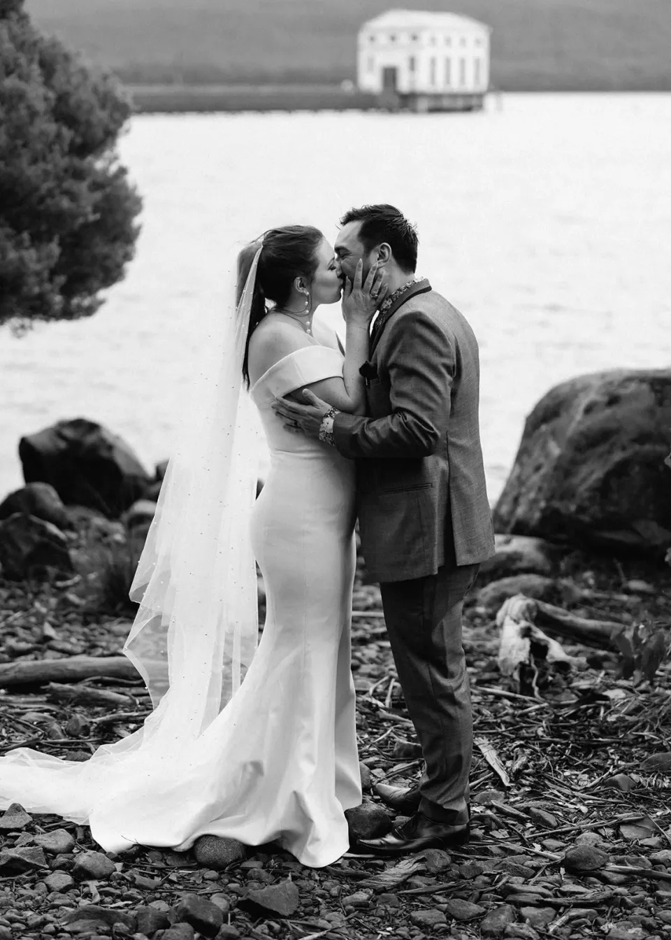 Black-and-white photograph of Sarah and Jono embracing beside the lake, her veil drifting behind her, at Pumphouse Point, Lake St Clair, Tasmania, for an Elopement Collective elopement. Photo by Bulb Creative.