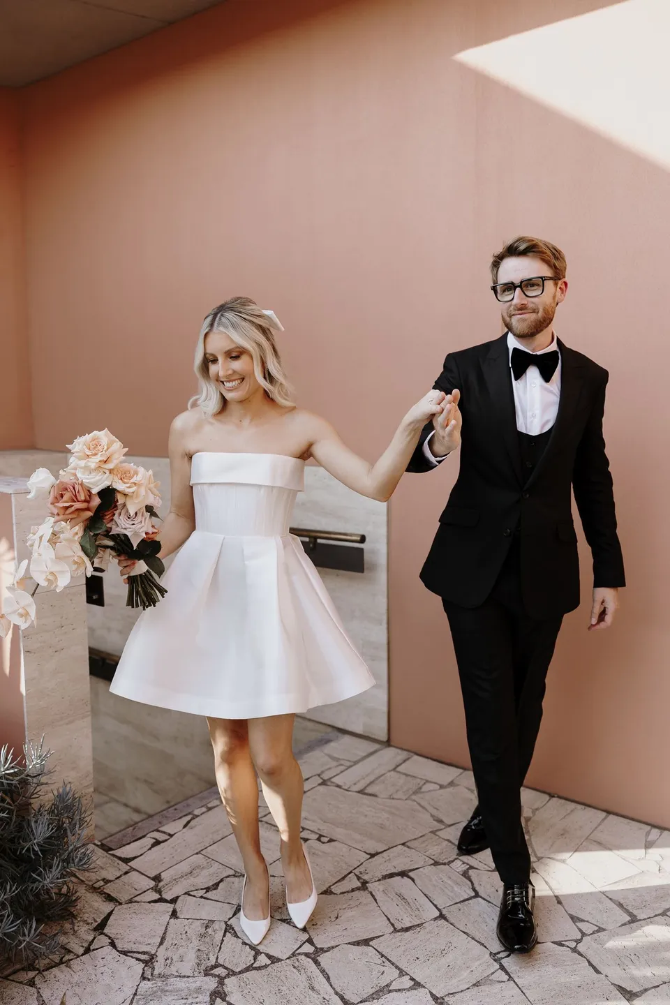Danielle and Adam walking hand in hand across the stone terrace beside a pink wall, at The Calile Hotel in Fortitude Valley, Brisbane, for an Elopement Collective elopement. Photo by George Bowden.
