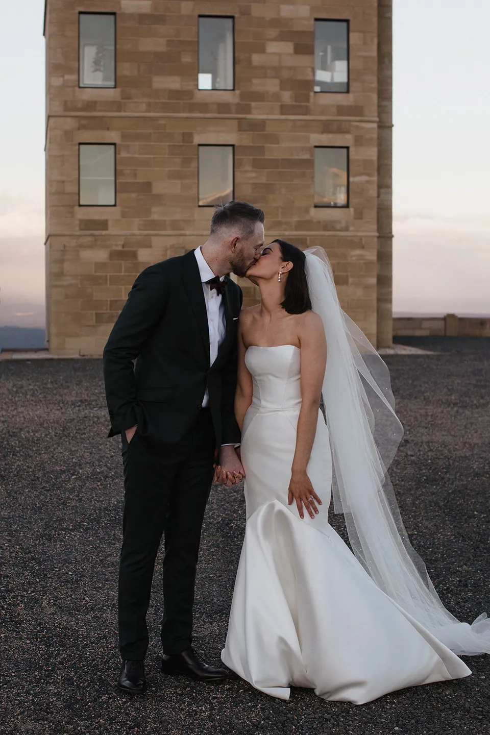 Meg and Ryan kissing in front of the tower, framed by sandstone walls and the evening sky. At The Keep on Tasmania’s east coast, for an Elopement Collective elopement. Photograph by Jenna Agius.