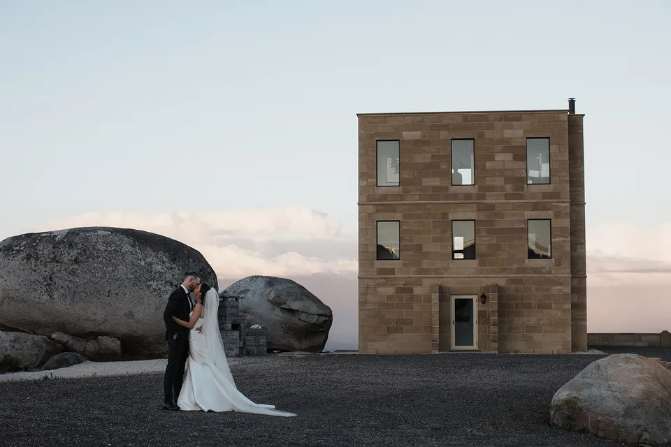 Meg and Ryan embracing between enormous granite boulders and the sandstone tower. At The Keep on Tasmania’s east coast, for an Elopement Collective elopement. Photograph by Jenna Agius.