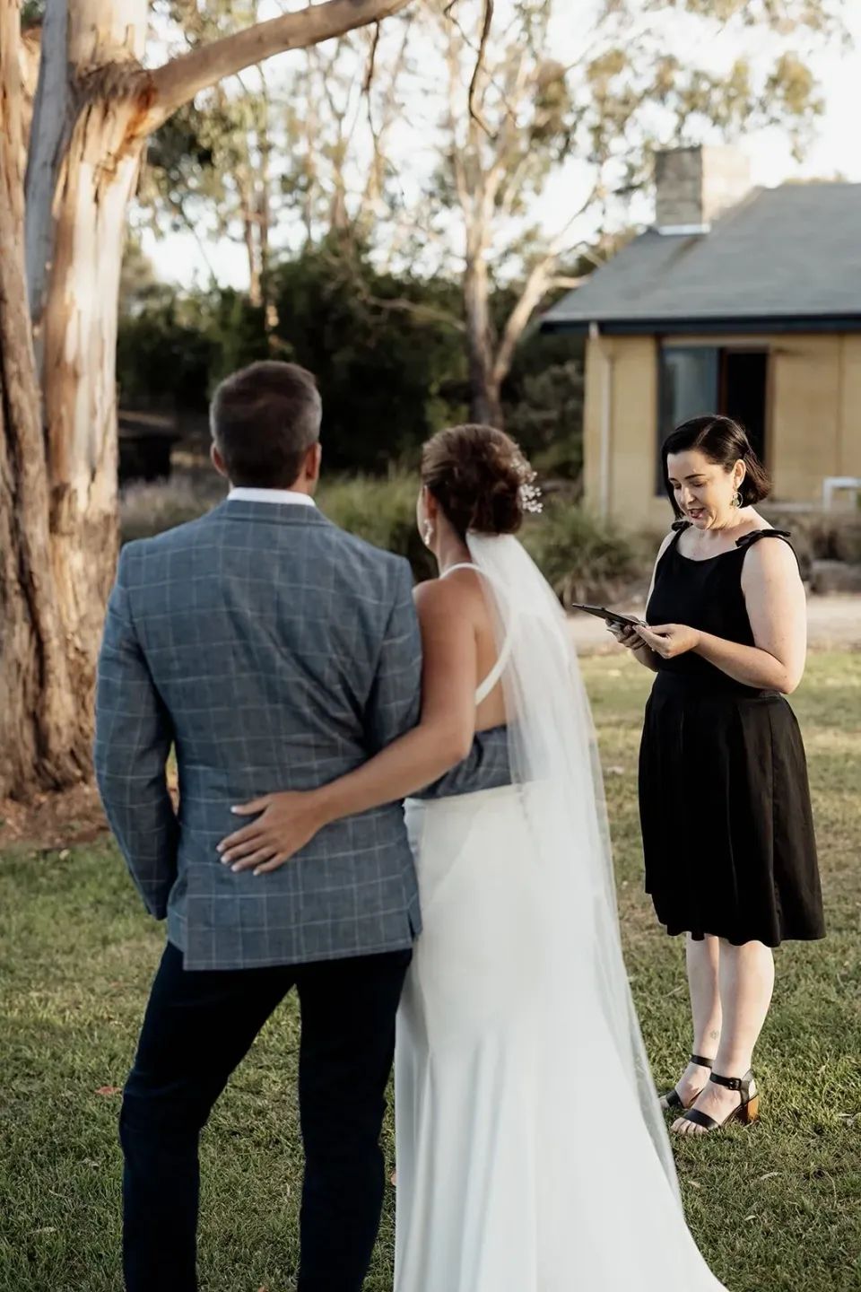 A couple stand with their celebrant on a lawn beside a cabin.