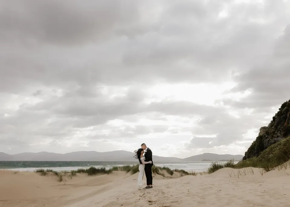 A couple embrace on a sandy beach beneath a cloudy sky, with dunes and cliffs nearby.