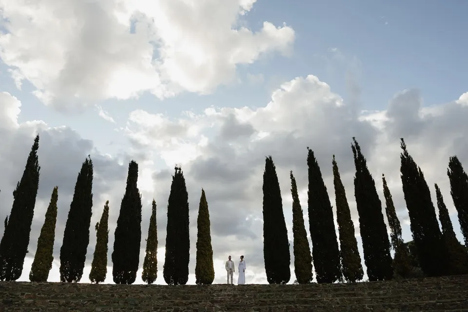 Renee and Mitch standing between tall cypress trees above the stone amphitheatre, beneath a wide, clouded sky. At Piermont Retreat on Tasmania’s east coast, for an Elopement Collective elopement. Photograph by Bulb Creative.