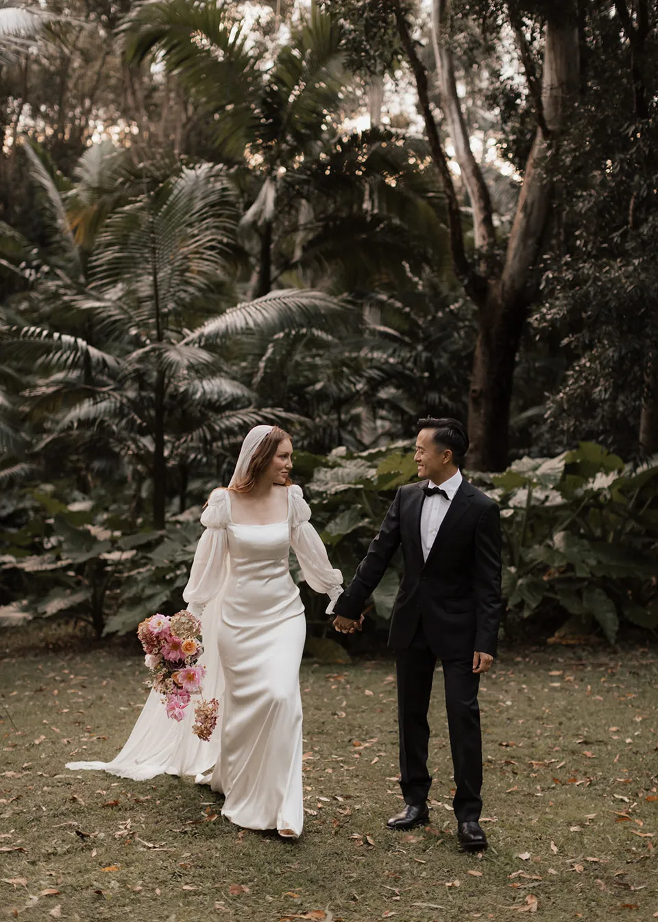 The couple holding hands beneath the tall palms, Elyse carrying her bouquet, with flowers by The Perfect Party Co. At Fig Tree Villa in Ewingsdale, Byron Bay, for an Elopement Collective elopement. Photograph by Bulb Creative.