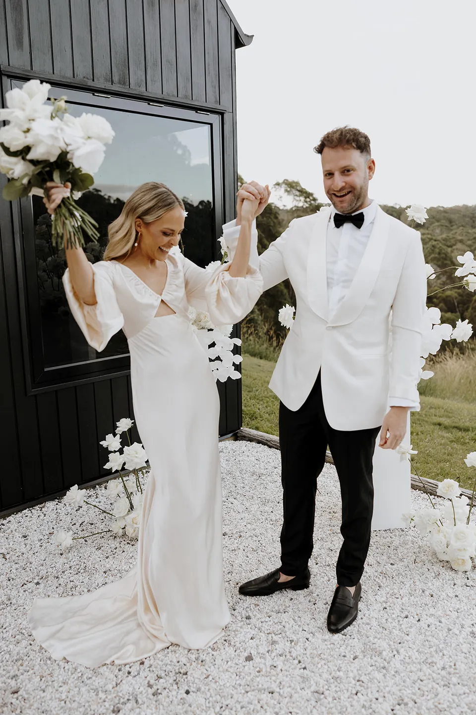 Gabrielle lifting her bouquet as Greg raises their joined hands in celebration outside the cabin, at Roundhill Retreat in Obi Obi, Sunshine Coast Hinterland, Queensland, for an Elopement Collective elopement. Photo by George Bowden.