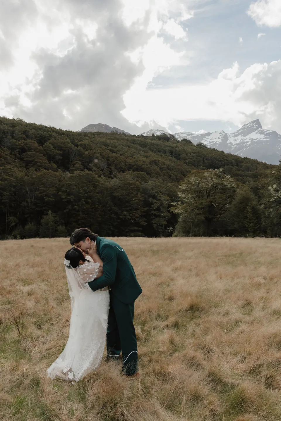 Jaeda and Adrian kissing in the open field after their ceremony, with forest and snowy mountains behind them, in the field on the road to Paradise near Glenorchy, in the Queenstown region of New Zealand, for an Elopement Collective Queenstown elopement. Photograph by Bulb Creative.
