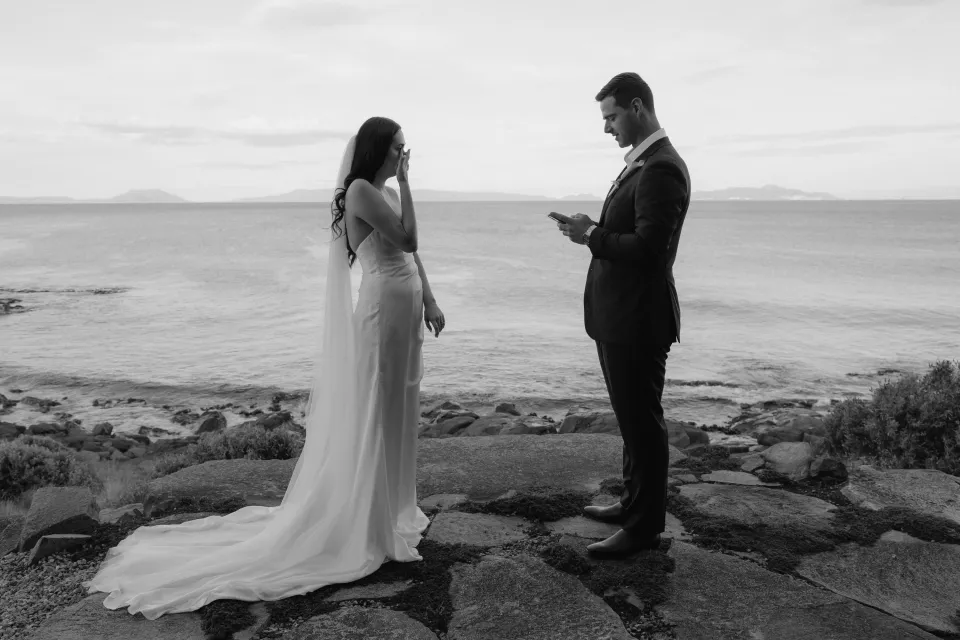 Black and white photograph of Anthony reading his vows from his phone to Dani, with her veil trailing over the rocks, at Thalia Haven on Tasmania’s east coast, for an Elopement Collective elopement. Photograph by Bulb Creative.