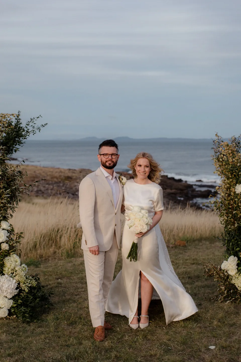 Jemmah and Jack standing together with her bouquet by The Romantics, at Thalia Haven on the East Coast of Tasmania, Australia, for an Elopement Collective elopement. Photograph by Joey Willis.