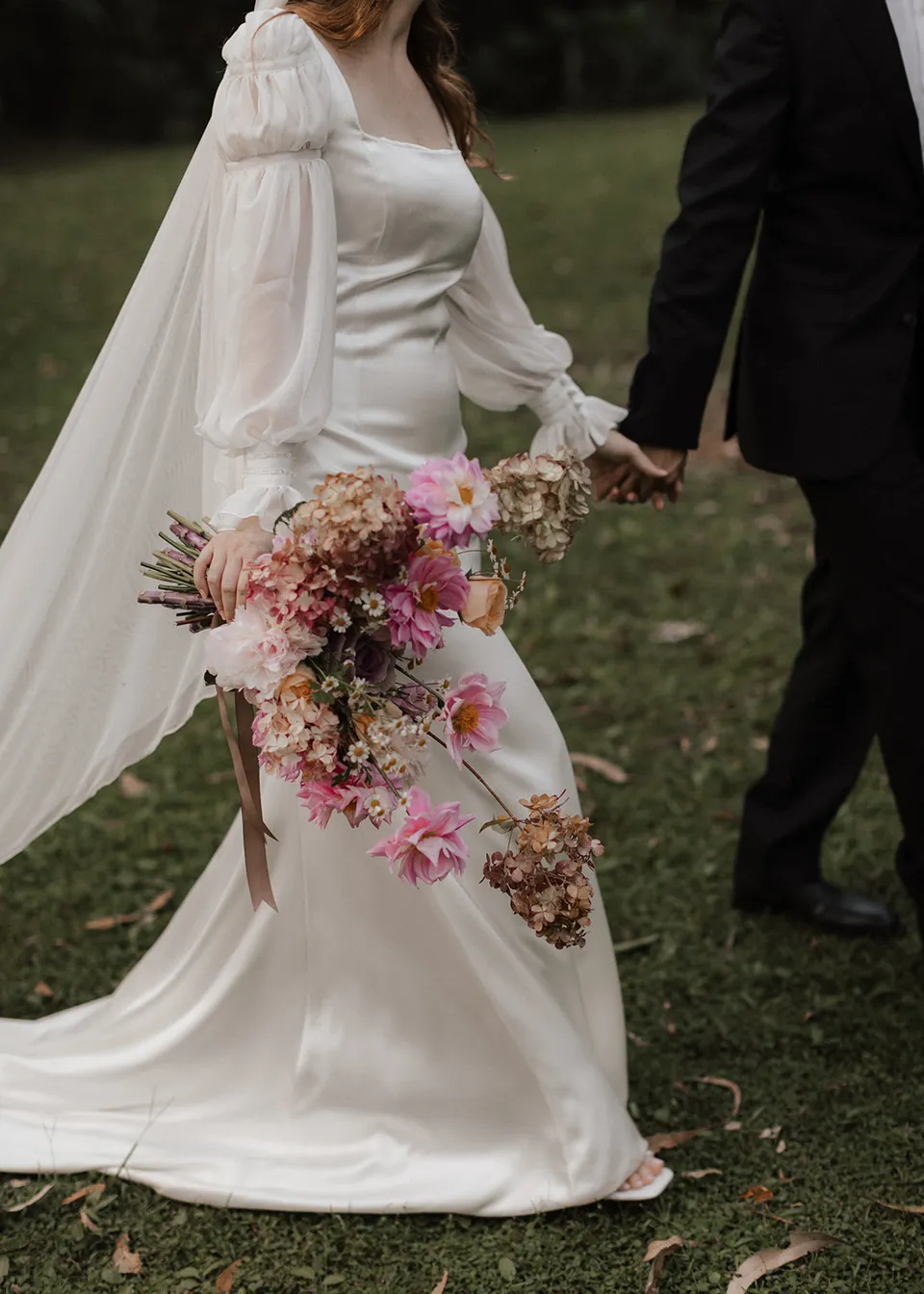 Close-up of Elyse's bouquet and flowing veil as the couple walk hand in hand, with flowers by The Perfect Party Co. At Fig Tree Villa in Ewingsdale, Byron Bay, for an Elopement Collective elopement. Photograph by Bulb Creative.