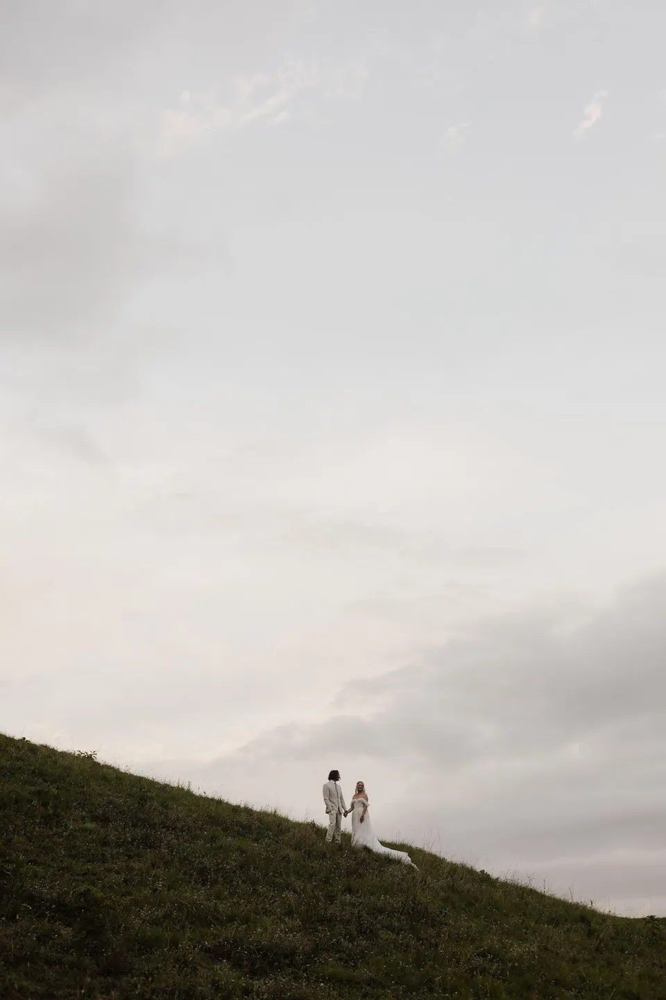 Distant view of Bridget and James together on the crest of a grassy hill. At Cudgera Creek in northern New South Wales, for an Elopement Collective elopement. Photograph by Bulb Creative.