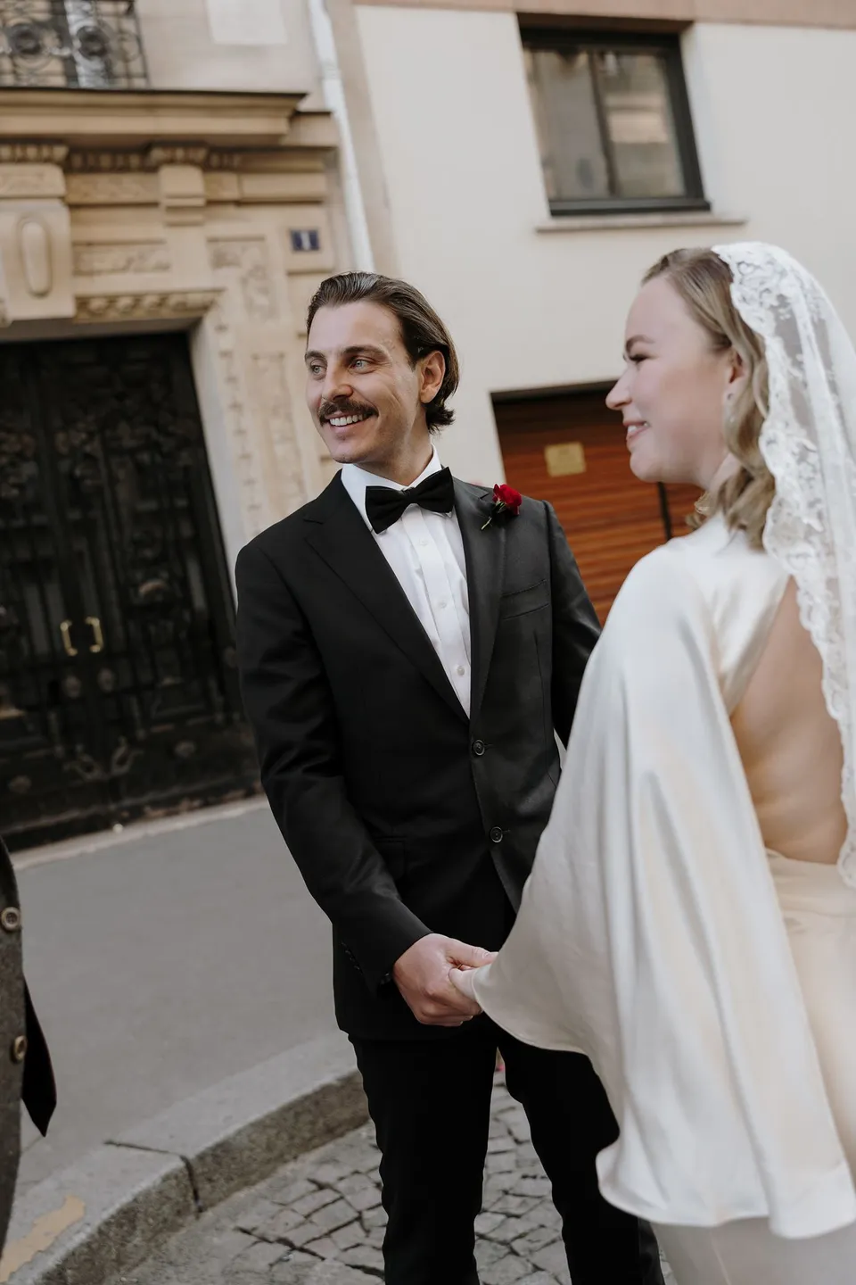 Samuel smiling in his black tuxedo beside Kara in her lace veil during their ceremony, at Avenue de Camoëns in Paris, France, during an Elopement Collective elopement. Photograph by George Bowden.
