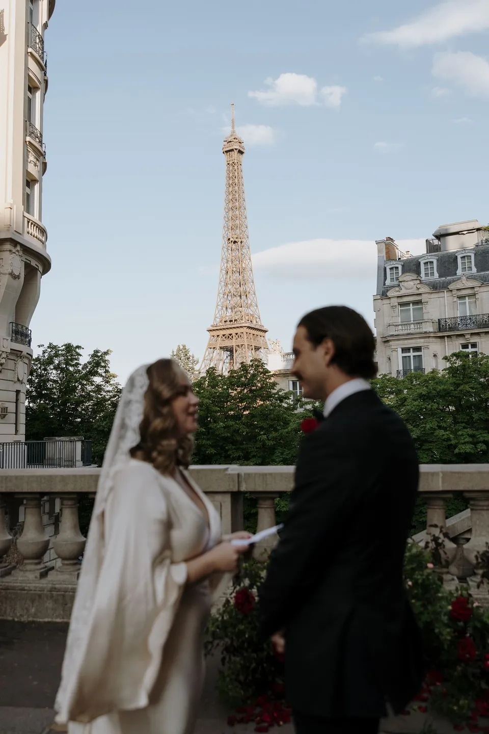 Kara and Samuel holding hands and facing each other during their ceremony, with Ivy Bleu’s red flowers and the Eiffel Tower behind them, at Avenue de Camoëns in Paris, France, during an Elopement Collective elopement. Photograph by George Bowden.