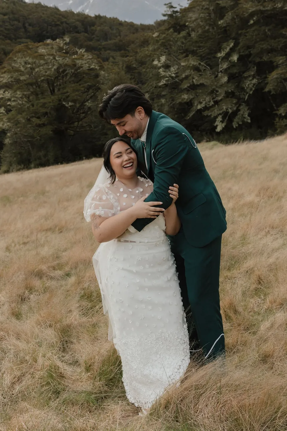 Jaeda laughing as Adrian kisses her cheek and holds her close in the golden grass, in the field on the road to Paradise near Glenorchy, in the Queenstown region of New Zealand, for an Elopement Collective Queenstown elopement. Photograph by Bulb Creative.