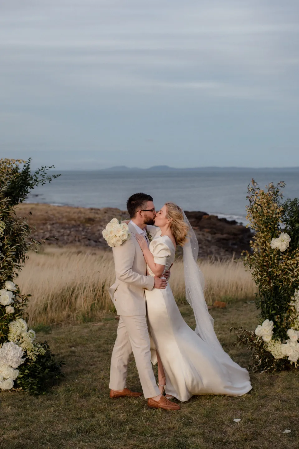 Jemmah and Jack kissing with her bouquet by The Romantics at her side, at Thalia Haven on the East Coast of Tasmania, Australia, for an Elopement Collective elopement. Photograph by Joey Willis.