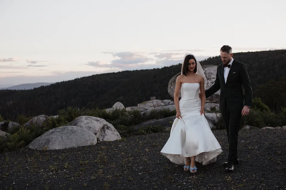 Meg lifting the hem of her dress as she and Ryan walk over gravel beside the granite boulders. At The Keep on Tasmania’s east coast, for an Elopement Collective elopement. Photograph by Jenna Agius.