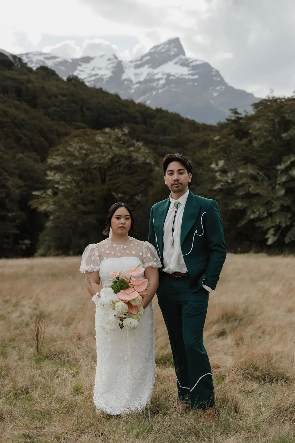 Jaeda and Adrian standing together beneath the mountains, with her pink and white bouquet by Orange Blossom Designs, in the field on the road to Paradise near Glenorchy, in the Queenstown region of New Zealand, for an Elopement Collective Queenstown elopement. Photograph by Bulb Creative.