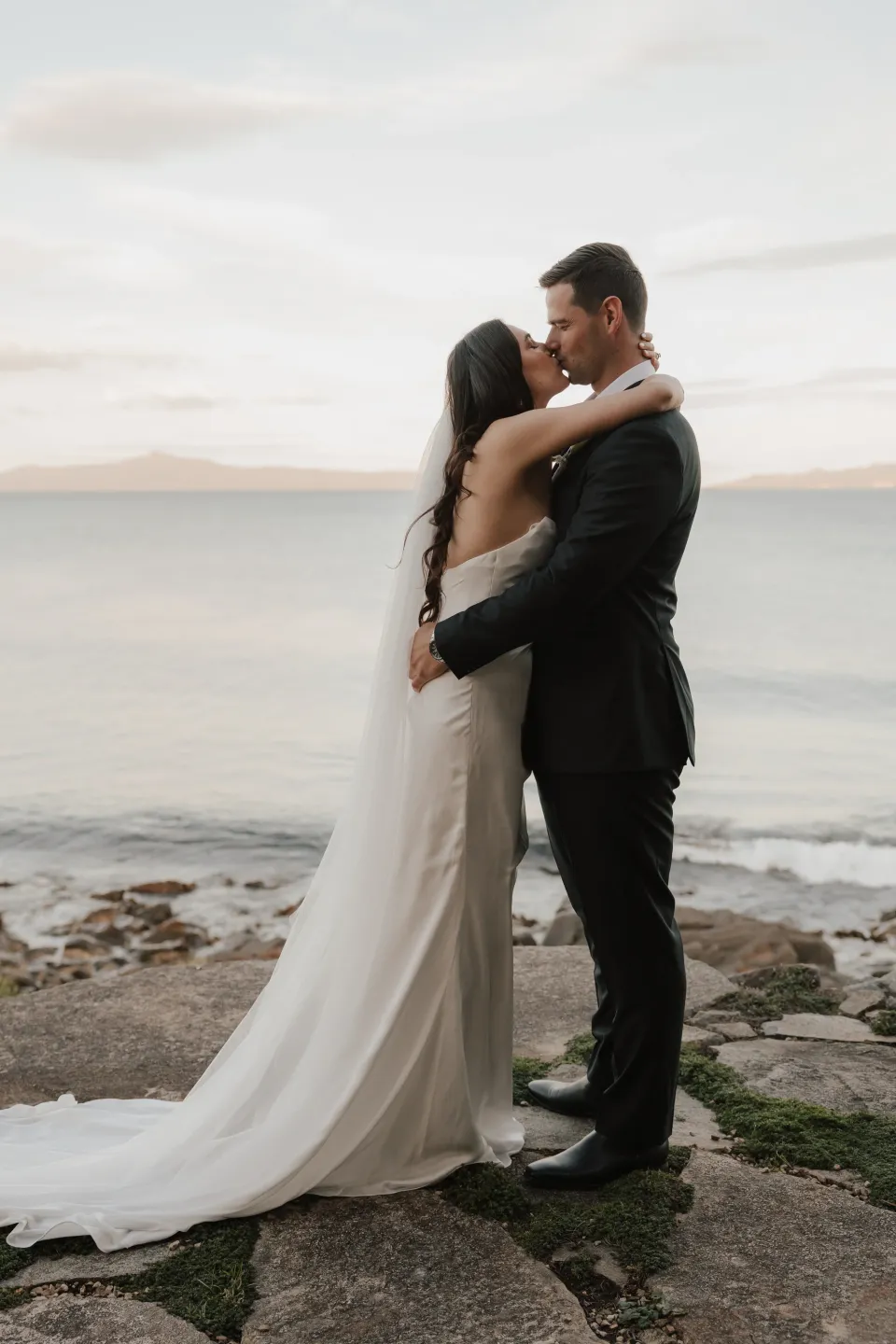 Dani and Anthony sharing a kiss on the rocks, with her white veil and dress flowing towards the bay, at Thalia Haven on Tasmania’s east coast, for an Elopement Collective elopement. Photograph by Bulb Creative.