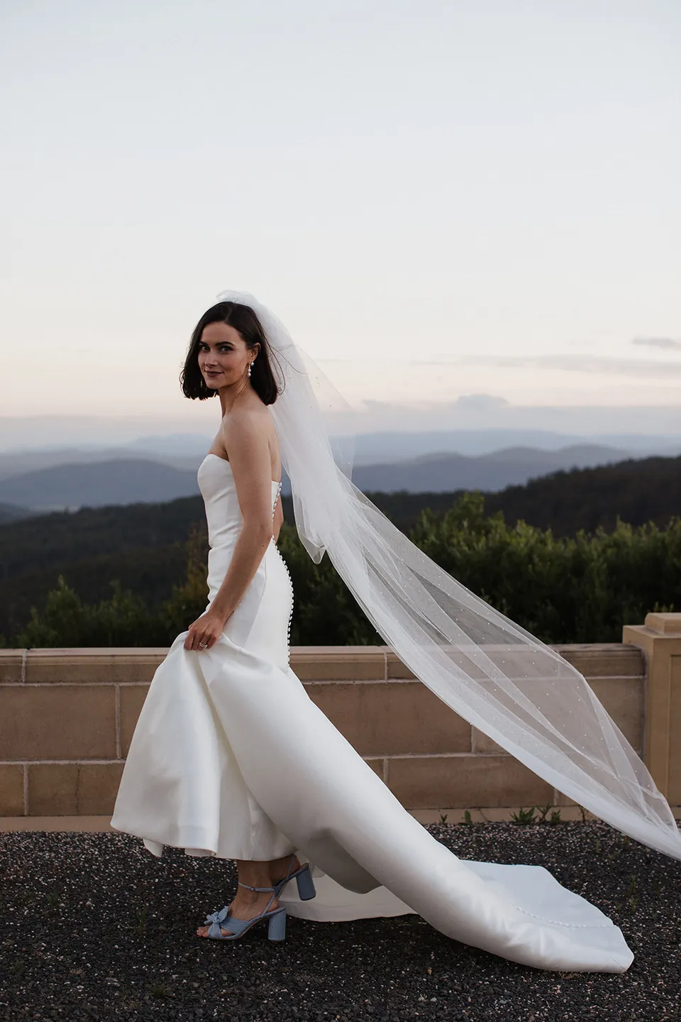 Meg looking back on the stone terrace, with her long veil trailing towards the hilltop view. At The Keep on Tasmania’s east coast, for an Elopement Collective elopement. Photograph by Jenna Agius.