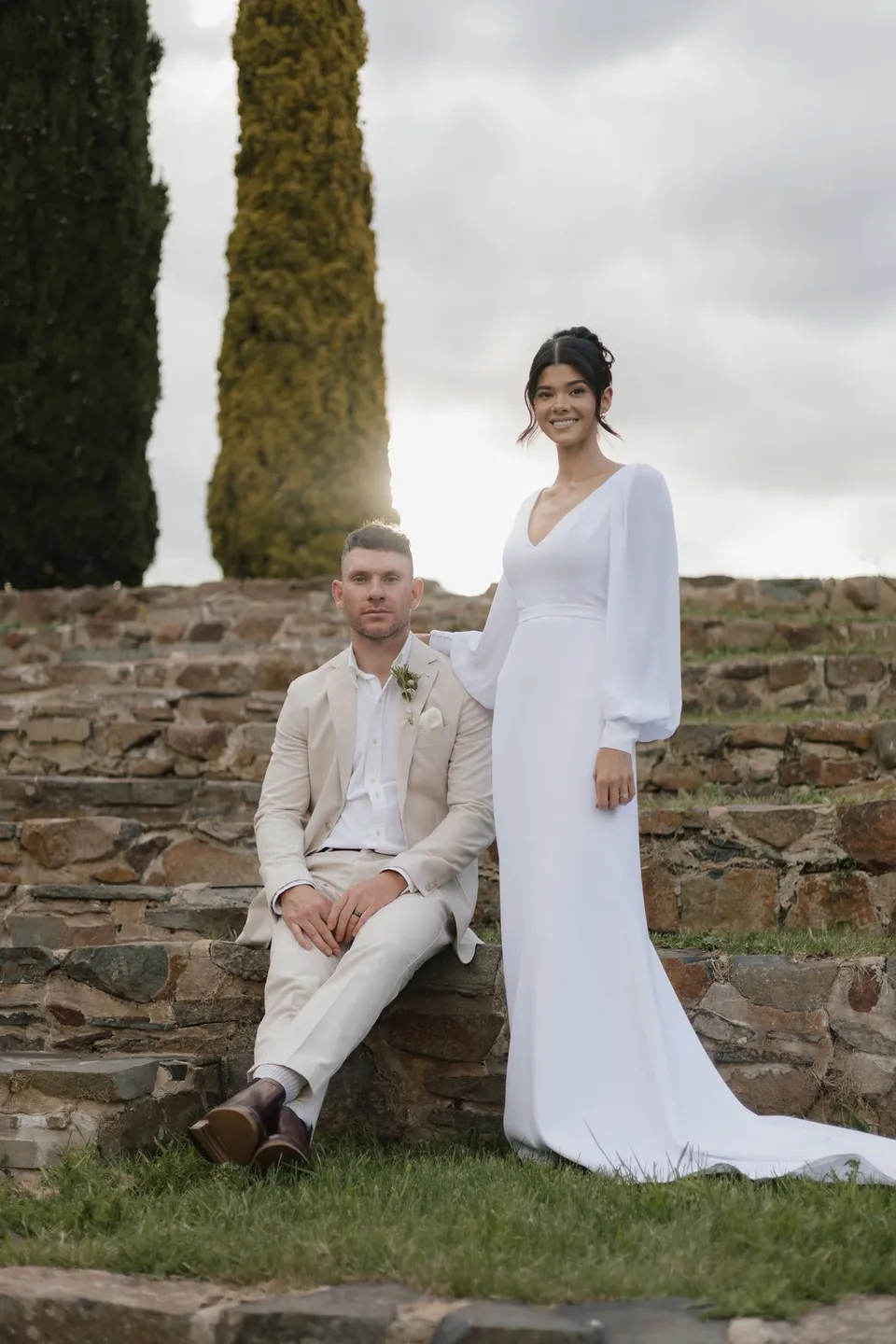 Mitch seated on the stone amphitheatre steps beside Renee, with tall cypress trees rising behind them. At Piermont Retreat on Tasmania’s east coast, for an Elopement Collective elopement. Photograph by Bulb Creative.