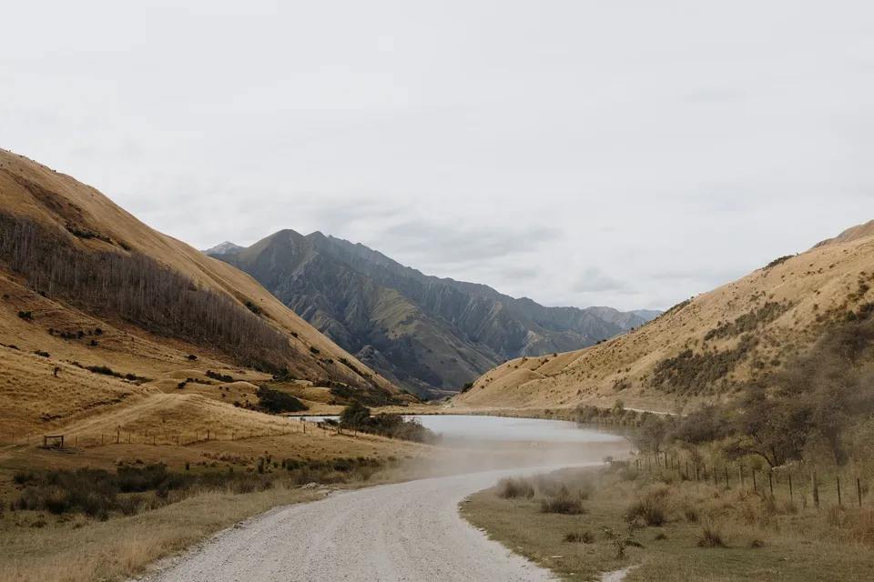 A gravel road winding beside a mountain lake and golden hills on the journey to the ceremony, in Queenstown, New Zealand, for an Elopement Collective Queenstown elopement. Photo by George Bowden.