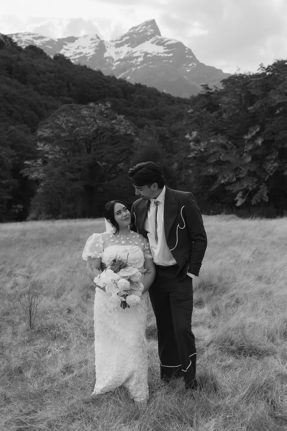 A black and white portrait of Jaeda and Adrian smiling together in the meadow, with her Orange Blossom Designs bouquet, in the field on the road to Paradise near Glenorchy, in the Queenstown region of New Zealand, for an Elopement Collective Queenstown elopement. Photograph by Bulb Creative.
