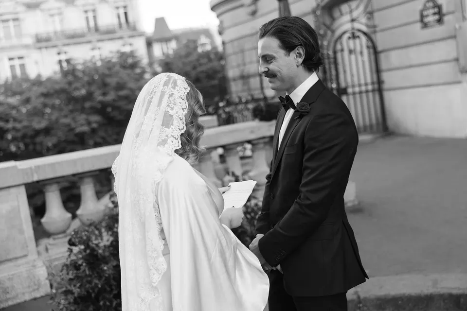 Black and white photograph of Kara and Samuel holding hands during their ceremony, with Samuel smiling towards her, at Avenue de Camoëns in Paris, France, during an Elopement Collective elopement. Photograph by George Bowden.