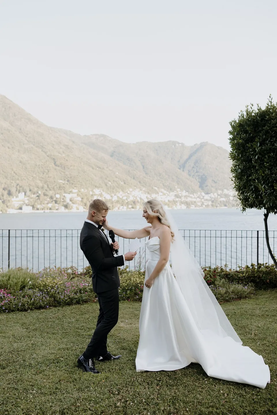 Zoe reaching up to Charlton’s face as he reads his vows, her long white train spread across the grass, at Passalacqua in Moltrasio, Lake Como, Italy, for an Elopement Collective elopement. Photo by George Bowden.