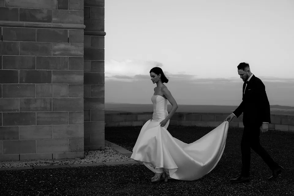 Black and white photograph of Meg walking past the tower as Ryan follows holding her dress train. At The Keep on Tasmania’s east coast, for an Elopement Collective elopement. Photograph by Jenna Agius.