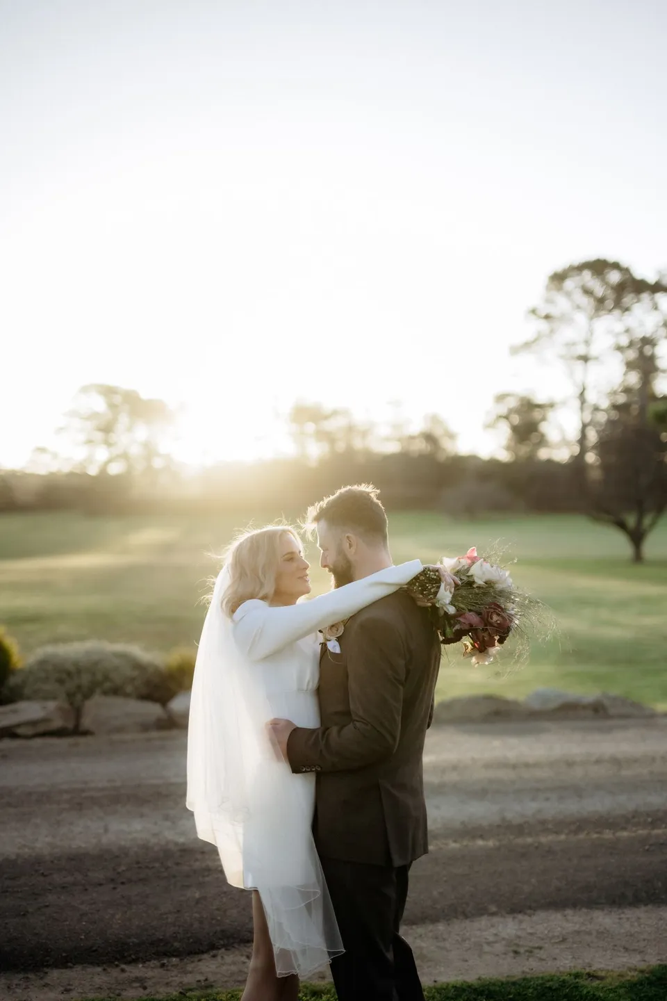 Kaitlyn and Russell embracing in low golden light, with her bouquet by The Romantics over his shoulder. At Piermont Retreat on Tasmania’s east coast, for an Elopement Collective elopement. Photograph by Omar of By The Wilde.