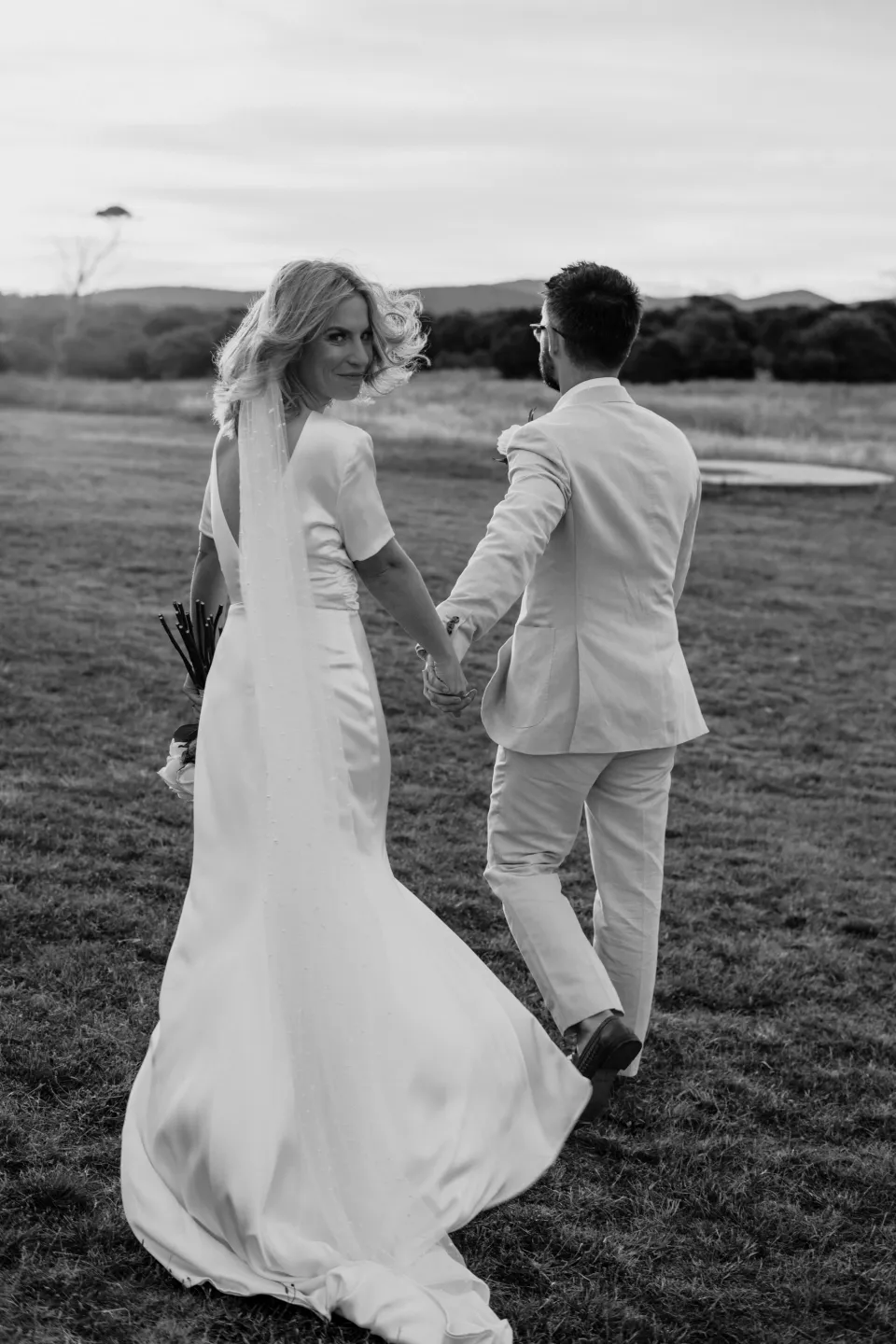 Black and white photograph of Jemmah turning towards the camera as they walk, at Thalia Haven on the East Coast of Tasmania, Australia, for an Elopement Collective elopement. Photograph by Joey Willis.