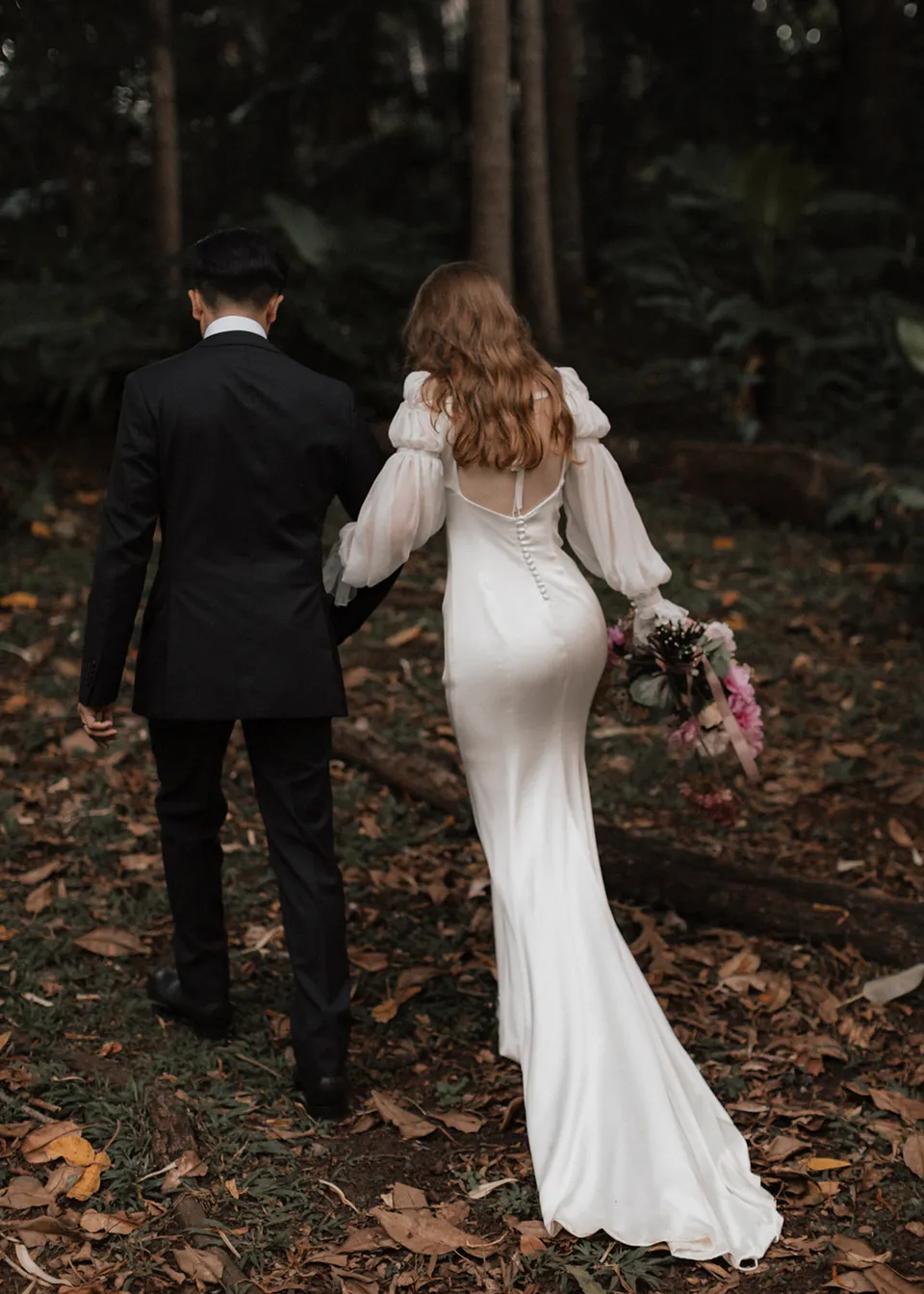 Rear view of Elyse and Jonathan walking through fallen leaves. At Fig Tree Villa in Ewingsdale, Byron Bay, for an Elopement Collective elopement. Photograph by Bulb Creative.