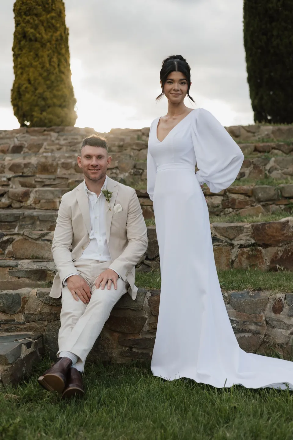 Renee smiling beside Mitch on the amphitheatre steps, with her white dress train resting on the grass. At Piermont Retreat on Tasmania’s east coast, for an Elopement Collective elopement. Photograph by Bulb Creative.