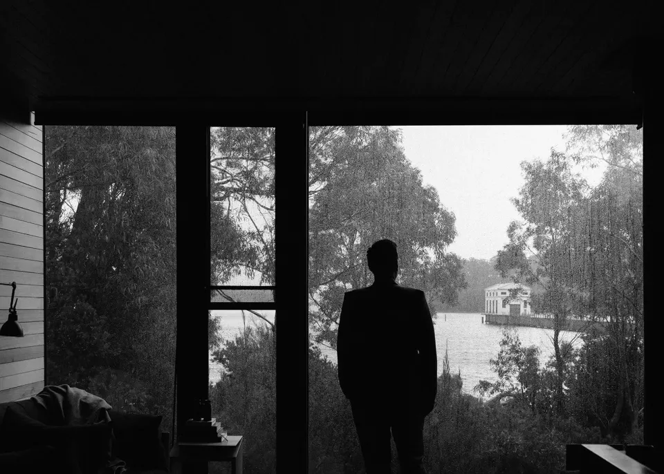 Black-and-white silhouette of Jono beside a tall window overlooking the lake and Pumphouse building, at Pumphouse Point, Lake St Clair, Tasmania, for an Elopement Collective elopement. Photo by Bulb Creative.