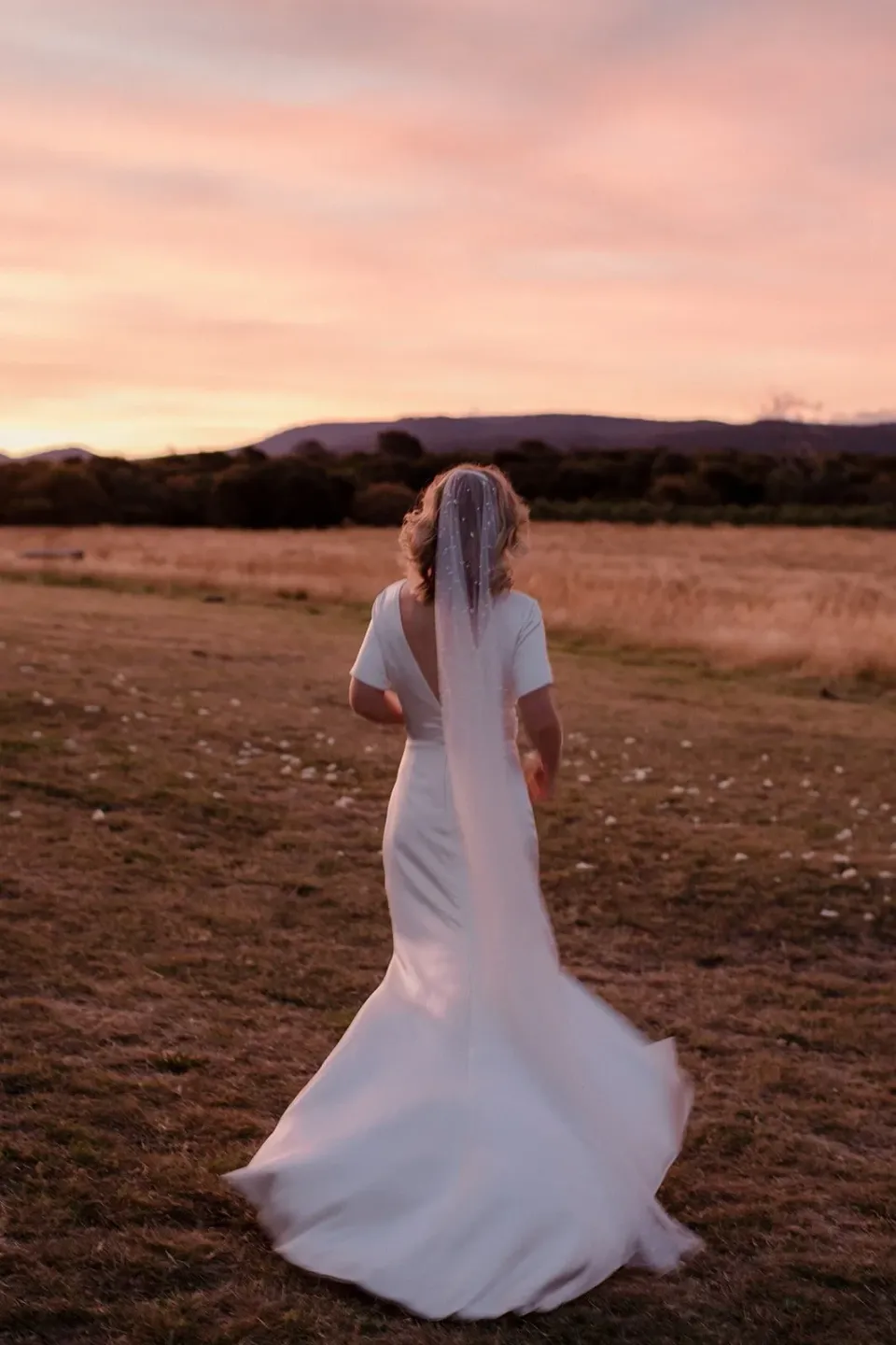 A bride stands in a paddock beneath a pink sunset, her veil moving in the breeze.