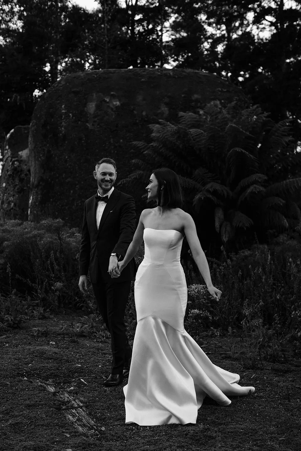 Black and white photograph of Meg and Ryan walking together through the woodland beside a fern-covered boulder. At The Keep on Tasmania’s east coast, for an Elopement Collective elopement. Photograph by Jenna Agius.
