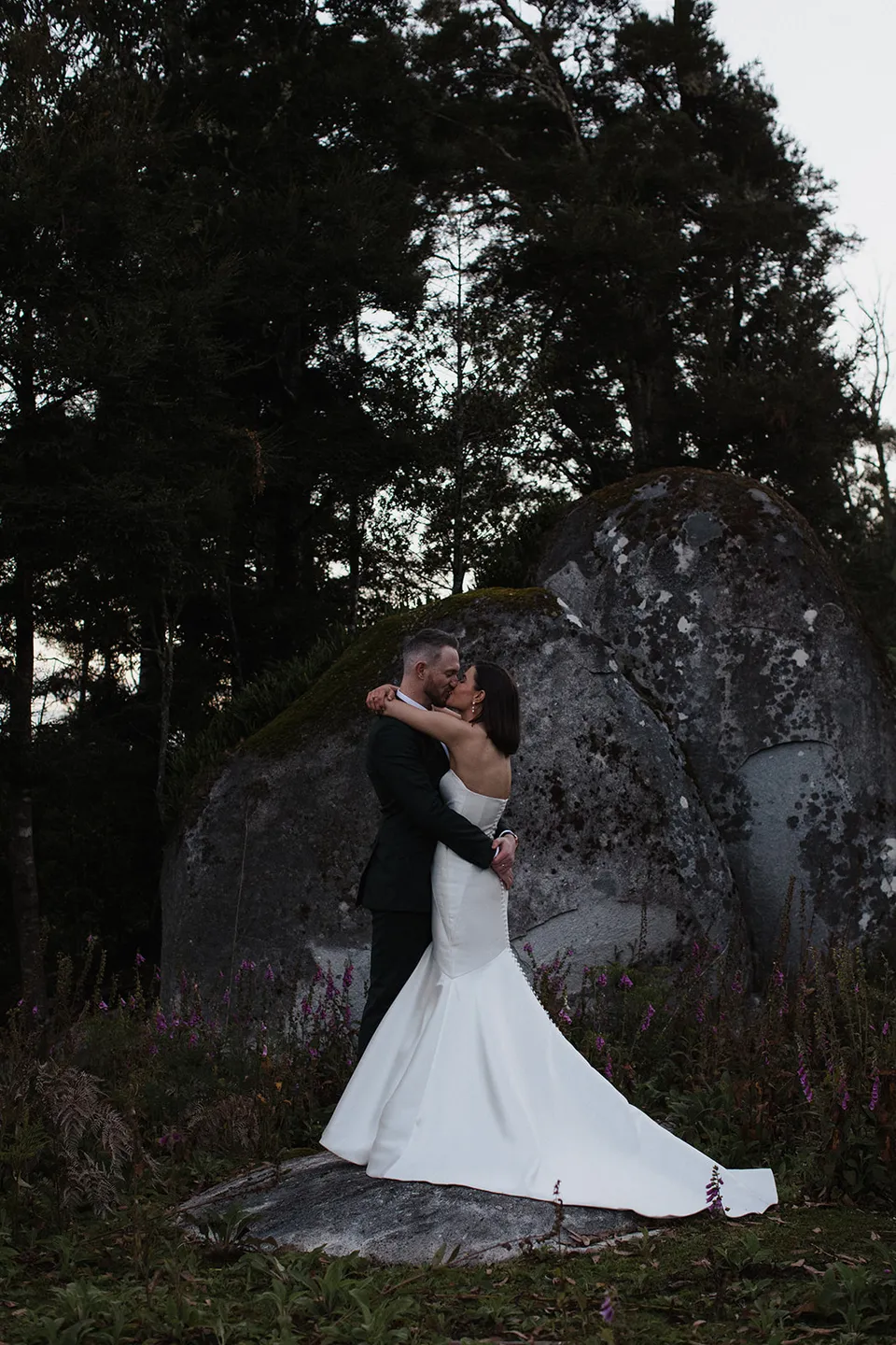 Meg and Ryan kissing beside a large boulder, with her dress spread over the rock and trees overhead. At The Keep on Tasmania’s east coast, for an Elopement Collective elopement. Photograph by Jenna Agius.