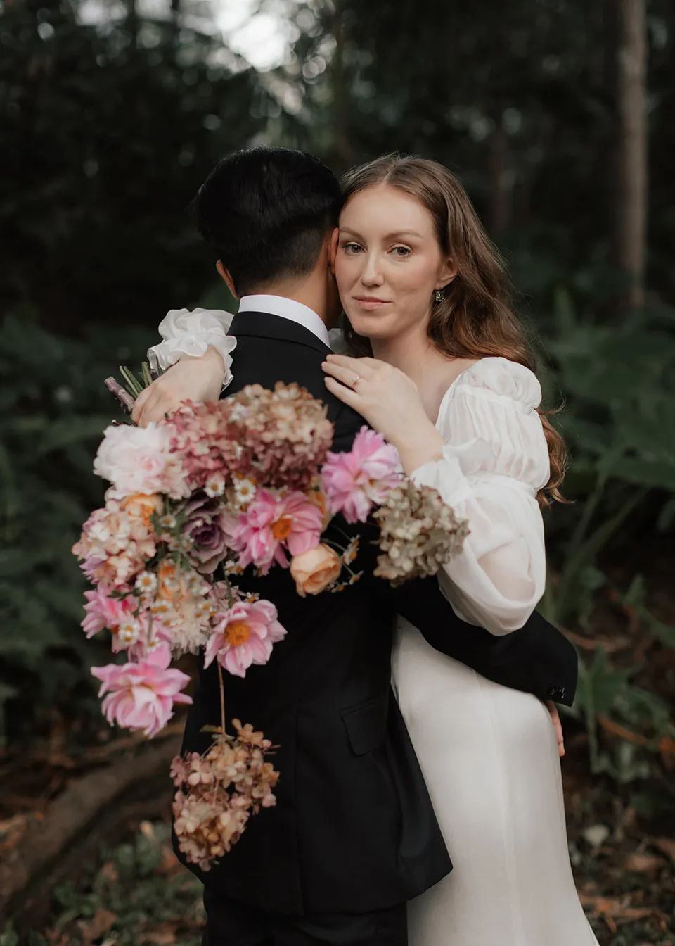 Elyse resting against Jonathan with her pink bouquet over his shoulder, with flowers by The Perfect Party Co. At Fig Tree Villa in Ewingsdale, Byron Bay, for an Elopement Collective elopement. Photograph by Bulb Creative.