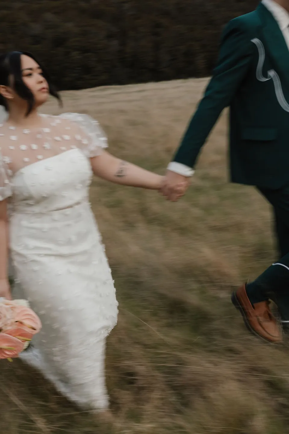 A motion-blurred photograph of Jaeda and Adrian running hand in hand through the golden grass, in the field on the road to Paradise near Glenorchy, in the Queenstown region of New Zealand, for an Elopement Collective Queenstown elopement. Photograph by Bulb Creative.
