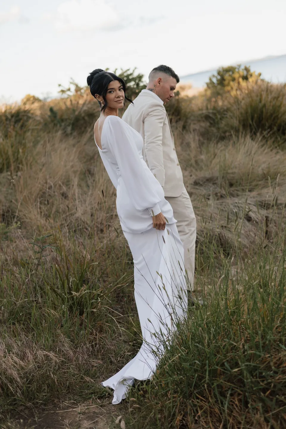 Renee looking back towards the camera as she and Mitch stand together among the tall coastal grasses. At Piermont Retreat on Tasmania’s east coast, for an Elopement Collective elopement. Photograph by Bulb Creative.