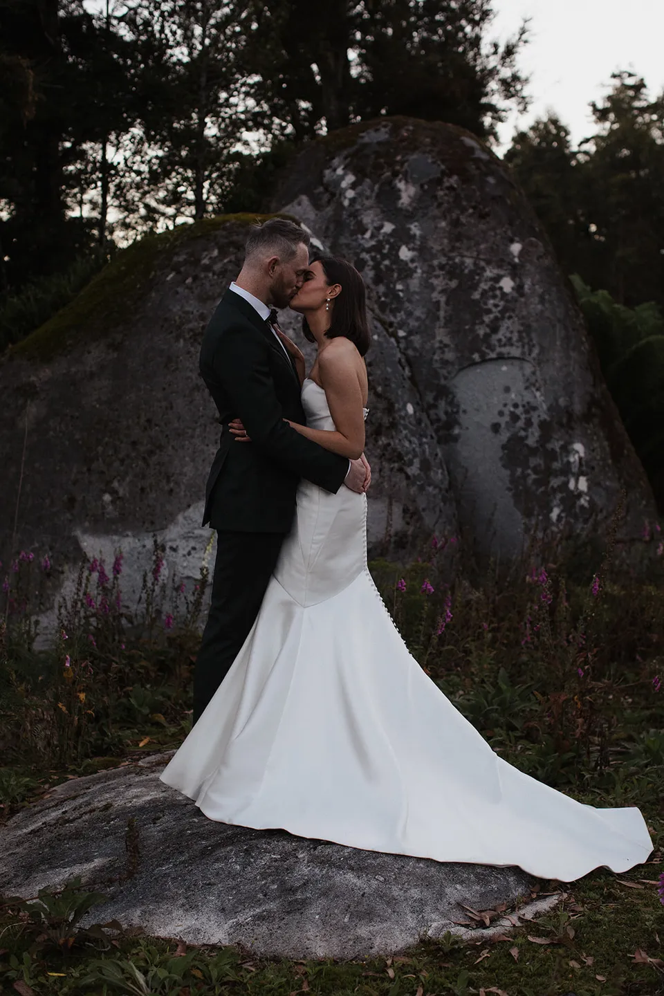 Meg and Ryan kissing on a rock beneath the trees, with the long train of her wedding dress flowing behind her. At The Keep on Tasmania’s east coast, for an Elopement Collective elopement. Photograph by Jenna Agius.