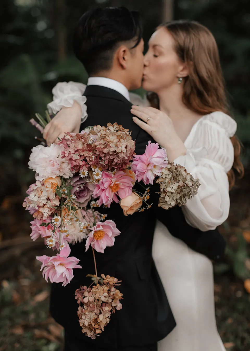 Elyse and Jonathan kissing beside her abundant pink and cream bouquet, with flowers by The Perfect Party Co. At Fig Tree Villa in Ewingsdale, Byron Bay, for an Elopement Collective elopement. Photograph by Bulb Creative.