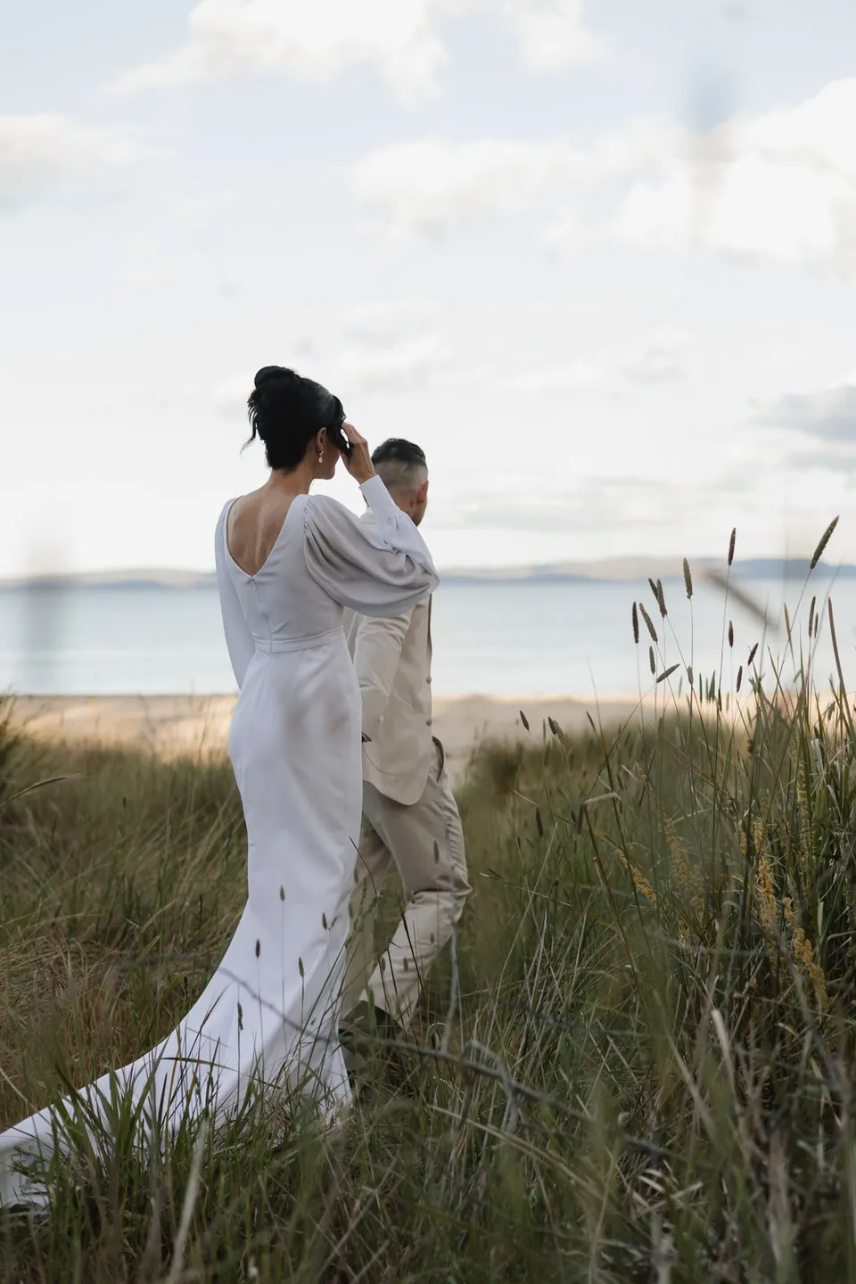 Renee and Mitch embracing among tall grasses above the water, with the open back and train of her dress visible. At Piermont Retreat on Tasmania’s east coast, for an Elopement Collective elopement. Photograph by Bulb Creative.