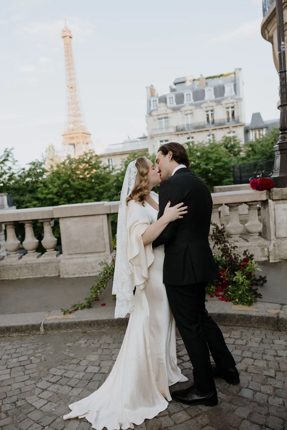 Kara and Samuel kissing beside Ivy Bleu’s red floral ceremony arrangements, with the Eiffel Tower behind them, at Avenue de Camoëns in Paris, France, during an Elopement Collective elopement. Photograph by George Bowden.