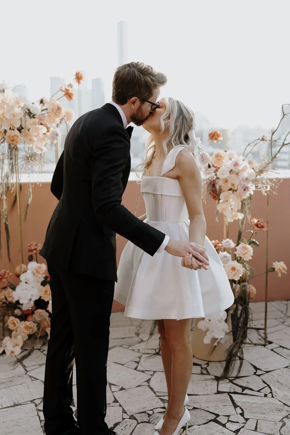 Danielle and Adam kissing on the terrace with peach and white flowers by Mrs Gibbons Florals behind them, at The Calile Hotel in Fortitude Valley, Brisbane, for an Elopement Collective elopement. Photo by George Bowden.