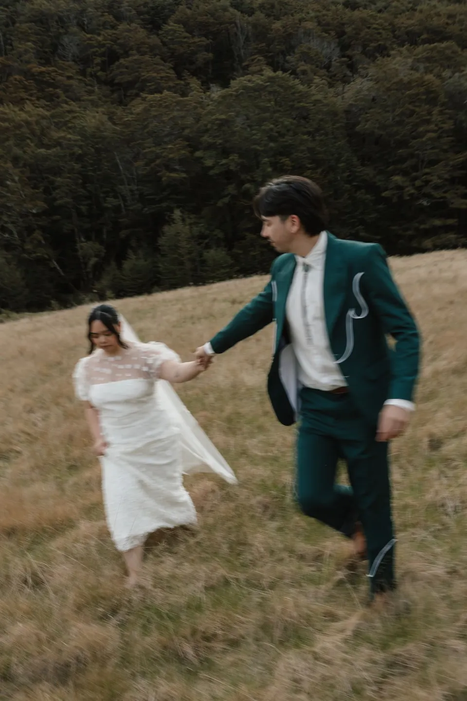Adrian leading Jaeda by the hand through the tall grass as she gathers her dress, in the field on the road to Paradise near Glenorchy, in the Queenstown region of New Zealand, for an Elopement Collective Queenstown elopement. Photograph by Bulb Creative.