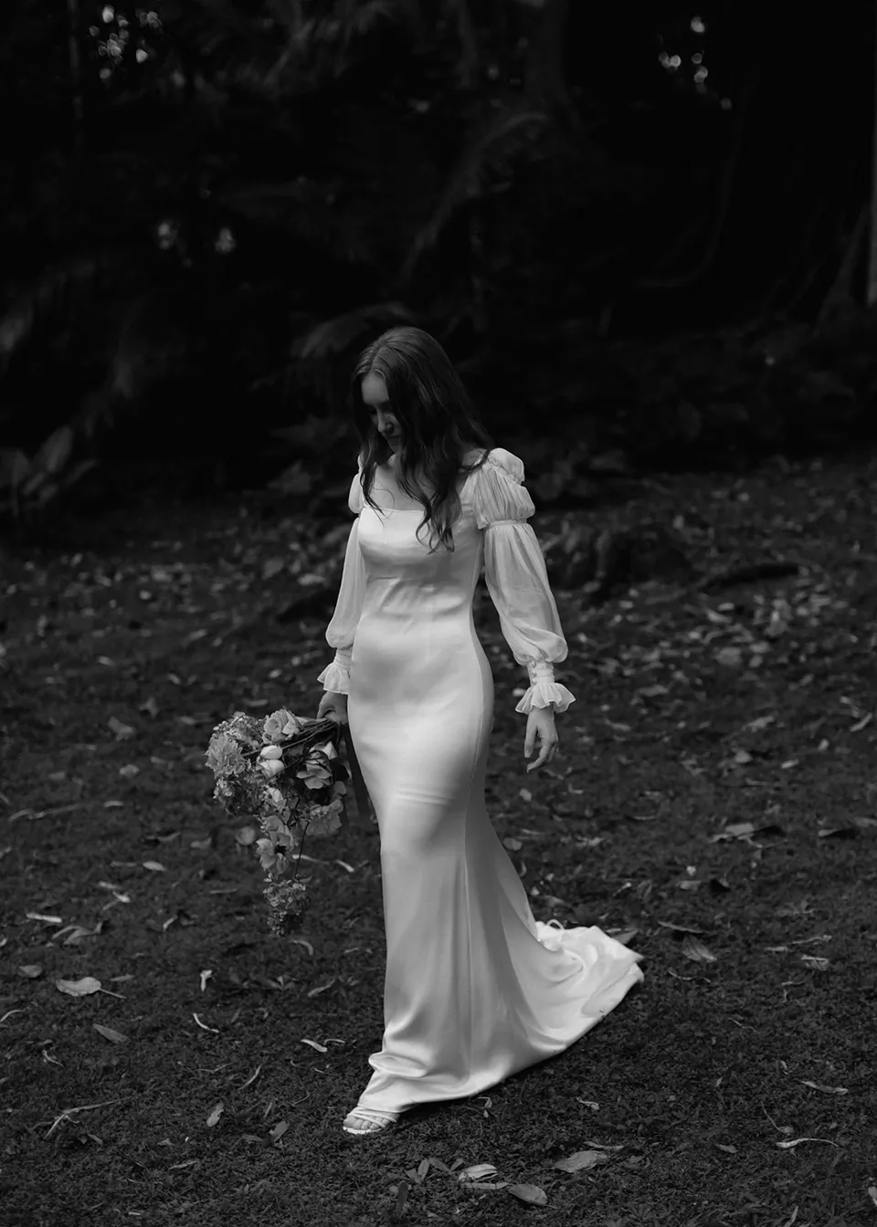 Black-and-white photograph of Elyse walking through the garden with her bouquet, with flowers by The Perfect Party Co. At Fig Tree Villa in Ewingsdale, Byron Bay, for an Elopement Collective elopement. Photograph by Bulb Creative.